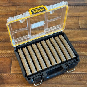 May include: A black and clear plastic storage case with a yellow accent and the text "TOUGHSYSTEM". The case is open, revealing nine tan-colored objects. The case has a handle and metal latches.
