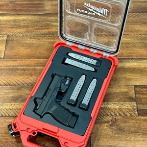 May include: Red and black hard case with a clear lid, containing a black handgun, three magazines, and a light. The case is open, revealing the contents. The brand name "Milwaukee" is visible on the lid.