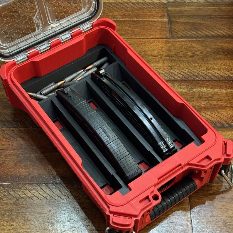 May include: A red, open, hard-shell case with a clear lid, containing black storage compartments. The compartments hold ammunition and black magazines. The case is on a wooden surface.