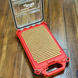 May include: A red, open, rectangular storage case filled with numerous brass-colored objects. The case has a clear lid with the brand name "Milwaukee" visible. The case is on a wooden surface.