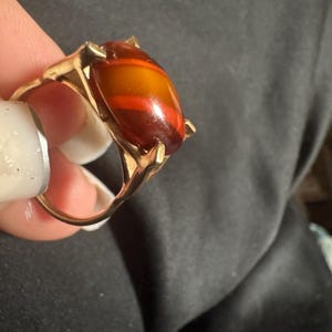 Könnte beinhalten: Ein goldfarbener Ring mit einem glatten, ovalen Cabochon-Stein. Der Stein zeigt horizontale Streifen in tiefem Bernstein, Orange und Dunkelbraun. Der Metallring ist schlicht gehalten und hält den Stein mit vier ausgeprägten Krappen fest.