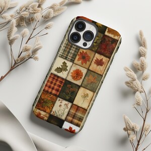 以下が含まれることがあります： 秋をテーマにした正方形のパッチワークデザインが施されたスマートフォンケース。様々な落ち葉、オレンジ色の花、チェック柄の布の質感が、茶色、オレンジ、緑、クリーム色の色合いで描かれており、3つのカメラレンズ用の切り抜きがあります。
