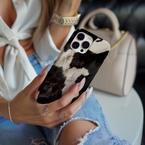 May include: A black and white cow print phone case for an iPhone. The case has a raised camera bump and a black and white cow print design.