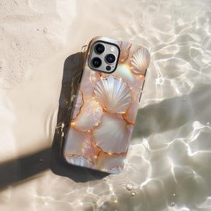 Puede incluir: Una funda de teléfono con diseño de conchas marinas y detalles dorados. La funda tiene un acabado nacarado con un patrón de conchas blancas contorneadas en dorado. La funda se muestra en agua, creando un efecto brillante.