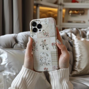 May include: A white iPhone case with a floral patchwork design. The case has a camera lens cutout and a white background with pink, green, and brown floral accents.