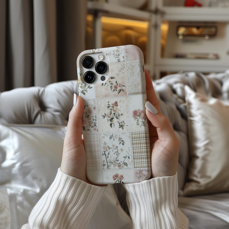 May include: A white iPhone case with a floral patchwork design. The case has a camera lens cutout and a white background with pink, green, and brown floral accents.