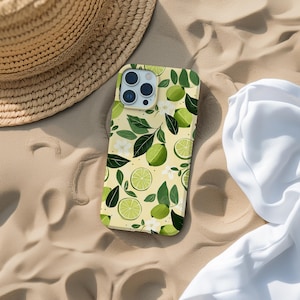 May include: A phone case with a yellow background featuring a pattern of green limes, leaves, and white flowers. The case is lying on sand next to a straw hat and a white cloth.
