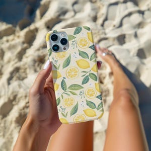 May include: A white phone case with a lemon and leaf pattern. The case has a camera lens cutout and a white background.