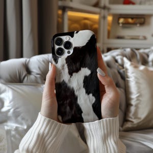 May include: A black and white cow print phone case for an iPhone. The case has a raised camera bump and a cutout for the charging port.
