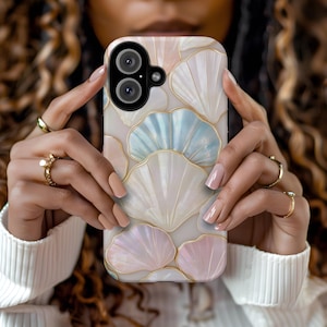 May include: A phone case featuring a seashell design in white, light blue, and pink, with gold-colored outlines. The case has a dual-lens camera cutout and is held by a person wearing rings.