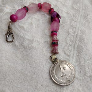 May include: A beaded keychain with a silver clasp and a coin pendant. The beads are various shades of pink and purple. The coin has Arabic script and a decorative edge.