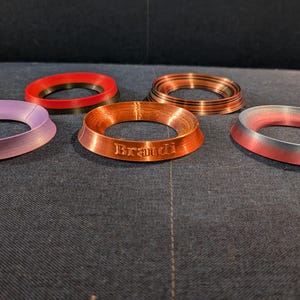 May include: Four different colored ring-shaped coasters. The coasters are made of a smooth, shiny material. One coaster is a light purple color, one is a dark red color, one is a copper color with the word "Brandi" engraved on it, and one is a light blue color with a red and silver gradient.