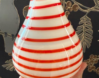 May include: A white glass vase with horizontal red stripes. The vase has a rounded bottom and tapers towards the top. The vase is held in a hand against a dark background with a bird and floral pattern.