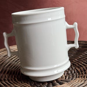 May include: A white ceramic mug with two curved handles. The mug has a cylindrical shape with a slightly flared top and a decorative band around the middle. The mug is sitting on a woven wicker surface.