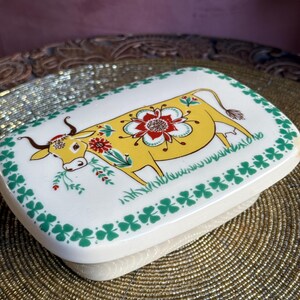 Op de afbeelding: Een rechthoekige botervloot met een geel koeienontwerp. De koe heeft een bloemenpatroon en is omgeven door groene klavers. De schaal is wit met een groene rand en een beige basis, ongeveer 15 cm lang.