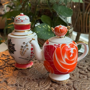 Könnte beinhalten: Zwei dekorative Keramik-Teekannen. Eine ist weiß mit roten und goldenen Akzenten und einem floralen Design. Die andere ist weiß mit einem roten und orangen Rosendesign. Beide haben goldene Verzierungen und kleine Griffe.