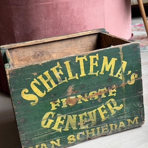 Op de afbeelding: Een vintage houten kist, geschilderd in groen met gele letters. De kist is gelabeld "Scheltema's Finste Genever Van Schiedam".