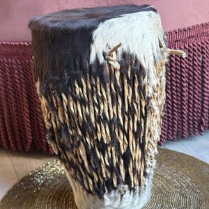 Pode incluir: Um tambor tradicional africano com uma cabeça de couro de vaca marrom e branca. O tambor é envolto com tiras tecidas de material marrom e tem uma franja de couro de vaca ao redor da parte inferior.