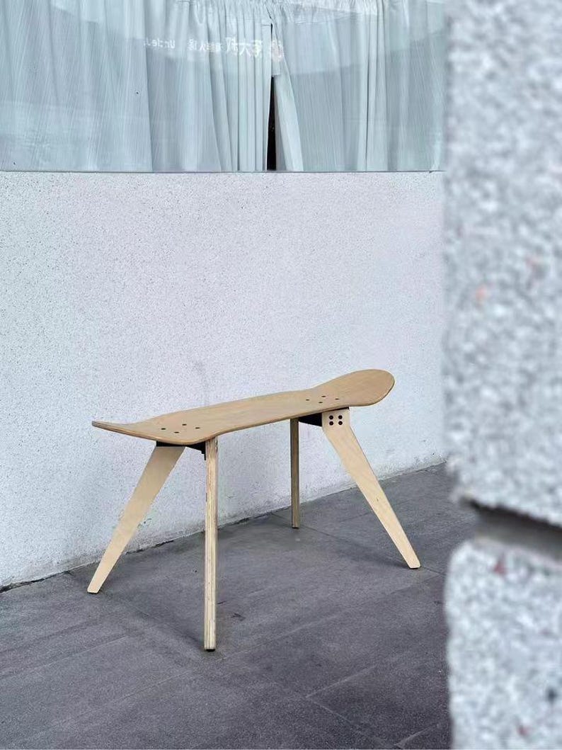K&ouml;nnte beinhalten: Eine Holzbank aus einem Skateboard-Deck mit vier Beinen. Die Bank ist hellbraun und hat eine nat&uuml;rliche Holzoptik. Die Bank steht auf einem grauen Betonboden.