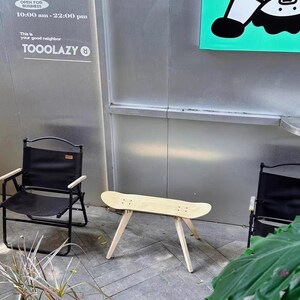K&ouml;nnte beinhalten: Ein Holztisch aus einem Skateboard mit vier Beinen, die wei&szlig; lackiert sind, steht auf einem grauen Fliesenboden. Zwei schwarze Klappst&uuml;hle stehen auf beiden Seiten des Tisches. Ein Schild mit dem Text "OPEN FOR BUSINESS 10:00 am - 22:00 pm This is your good neighbor TOOOLAZY" befindet sich an der Wand hinter dem Tisch. Ein gr&uuml;nes Schild mit einer Comicfigur befindet sich an der Wand hinter den St&uuml;hlen.