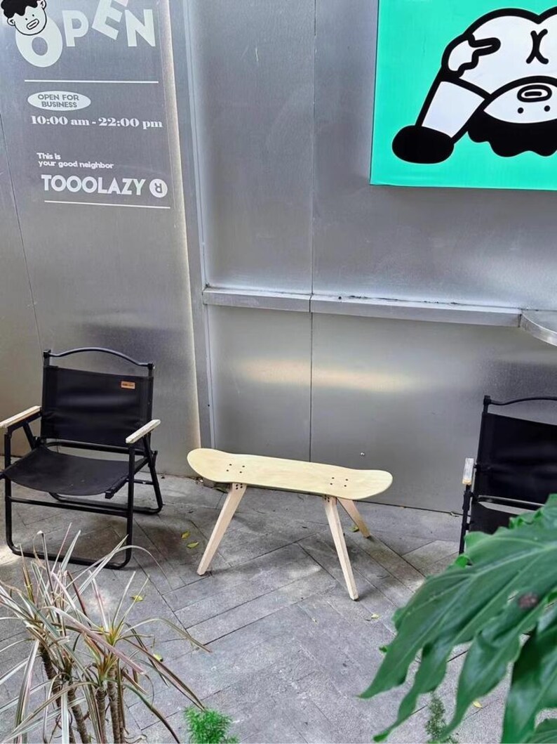 K&ouml;nnte beinhalten: Ein Holztisch aus einem Skateboard mit vier Beinen, die wei&szlig; lackiert sind, steht auf einem grauen Fliesenboden. Zwei schwarze Klappst&uuml;hle stehen auf beiden Seiten des Tisches. Ein Schild mit dem Text "OPEN FOR BUSINESS 10:00 am - 22:00 pm This is your good neighbor TOOOLAZY" befindet sich an der Wand hinter dem Tisch. Ein gr&uuml;nes Schild mit einer Comicfigur befindet sich an der Wand hinter den St&uuml;hlen.
