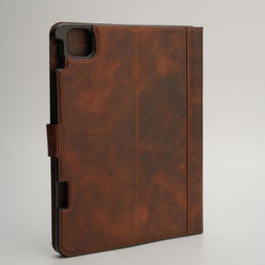 May include: Brown leather tablet case with a textured finish. The case has a black frame and a magnetic closure. The design includes a cutout for the camera and a vertical strap for securing the cover.