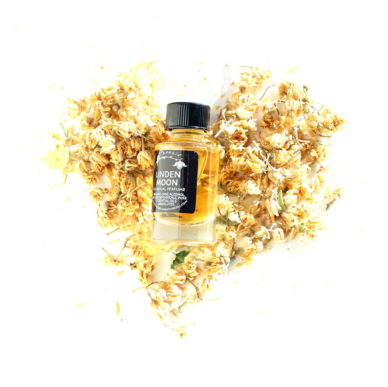 Peut inclure: Un petit flacon de parfum biologique Linden Moon en verre avec une &eacute;tiquette noire sur un fond blanc. Le flacon est entour&eacute; de fleurs jaunes s&eacute;ch&eacute;es.