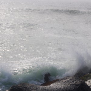 Pode incluir: Um close-up de uma costa rochosa com ondas quebrando. A &aacute;gua &eacute; de cor cinza claro e as rochas s&atilde;o de cor cinza escuro. As ondas criam um spray branco.