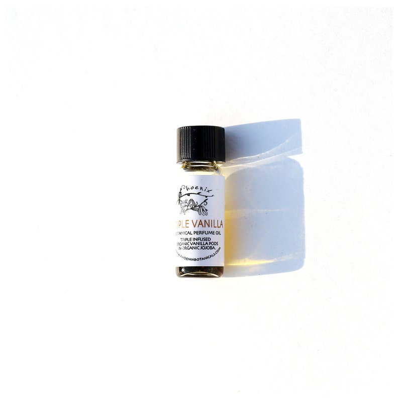 May include: A small glass bottle with a black cap containing a light brown liquid. The label reads "Triple Vanilla Botanical Perfume Oil Triple Infused Organic Vanilla Pods in Organic Jojoba" and features a small drawing of a tree branch with leaves.