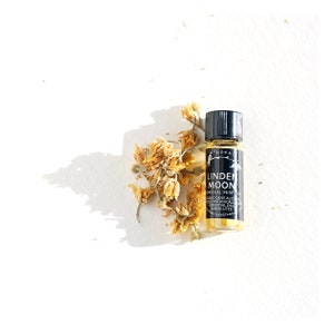 Pode incluir: Um pequeno frasco de vidro com uma tampa preta e um rótulo que diz "Linden Moon Botanical Perfume" com uma lista de ingredientes. O frasco está sobre uma superfície branca com flores amarelas secas.