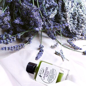 May include: A small glass bottle of lavender noir botanical perfume with a black cap. The bottle is sitting on a white surface with lavender flowers scattered around it. The label on the bottle reads "Phoenix Lavender Noir Botanical Perfume Organic Grape Sugarcane Pure Essential Oils Absolutes Alcohol, Infused Botanicals www.phoenxbotanicals.com"