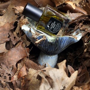 May include: A small glass bottle of lavender noir botanical perfume sits on a blue and white mushroom-shaped ceramic dish. The bottle has a black cap and a black label with white text that reads "Lavender Noir Botanical Perfume".
