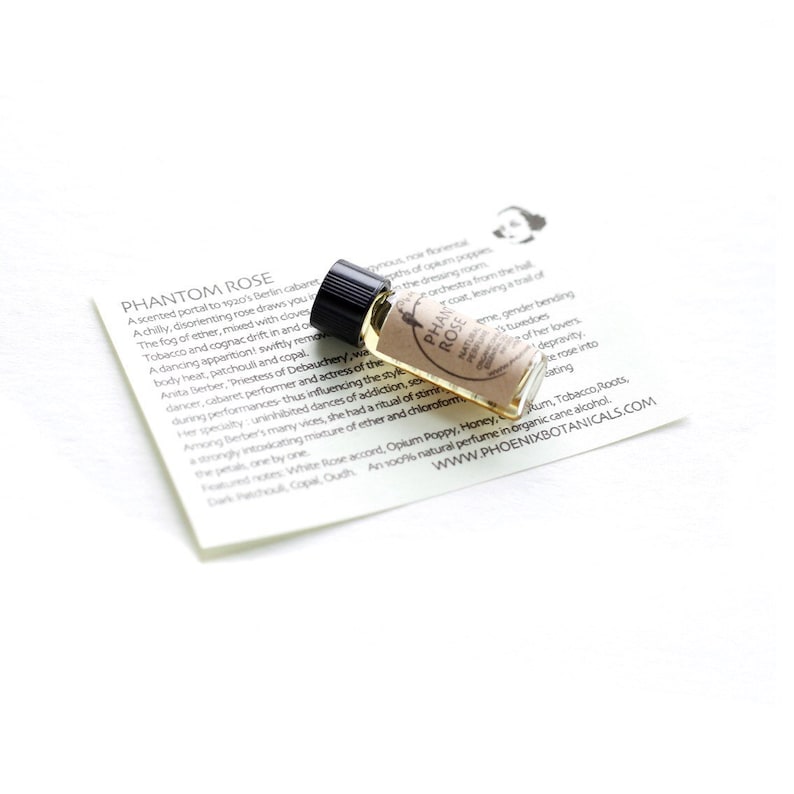 May include: A small glass bottle with a black cap containing a light brown liquid. The bottle is labeled "PHANTOM ROSE" and has a description of the scent, which includes notes of white rose, opium poppy, honey, rum, tobacco, roots, and oudh. The bottle is sitting on a white surface with a piece of paper that has text about the scent.