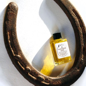 May include: A small glass bottle of Narcissus Cafe botanical perfume with a black cap. The bottle is filled with a yellow liquid and is sitting on a white surface next to a rusty horseshoe.