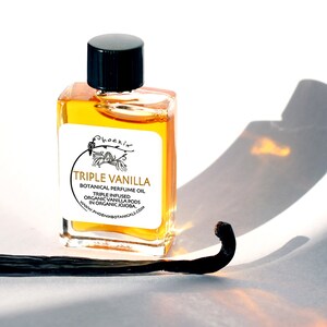 May include: A small glass bottle with a black cap filled with a golden liquid. The label reads "Phoenix Triple Vanilla Botanical Perfume Oil Triple Infused Organic Vanilla Pods in Organic Jojoba. www.phoenixbotanicals.com". A dried vanilla bean is in the foreground.
