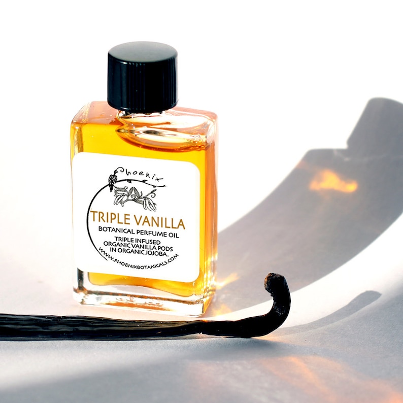 May include: A small glass bottle with a black cap filled with a golden liquid. The label reads "Phoenix Triple Vanilla Botanical Perfume Oil Triple Infused Organic Vanilla Pods in Organic Jojoba. www.phoenixbotanicals.com". A dried vanilla bean is in the foreground.