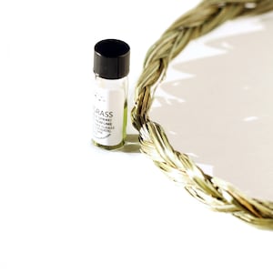 以下が含まれることがあります： 黒いキャップと「GRASS」のラベルが付いた小さなガラスの香水瓶。背景には、乾燥した草の編み込みリースがあります。香水瓶には、薄い緑色の液体が入っています。