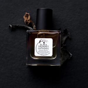 May include: A clear glass bottle with a black cap and a white label. The label reads "Phoenix Black Trumpet Chthonic Natural Perfume Organic Cane Alcohol Black Trumpet Mushroom Pure Plant Extracts Essential Oils www.phoenixbotanicals.com".