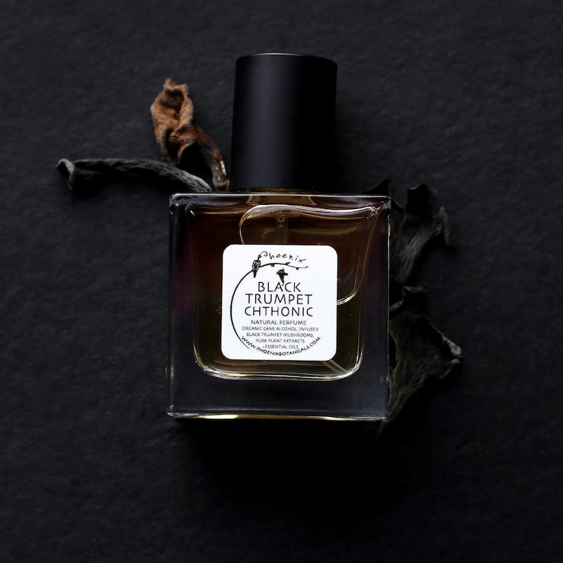 May include: A clear glass bottle with a black cap and a white label. The label reads "Phoenix Black Trumpet Chthonic Natural Perfume Organic Cane Alcohol Black Trumpet Mushroom Pure Plant Extracts Essential Oils www.phoenixbotanicals.com".