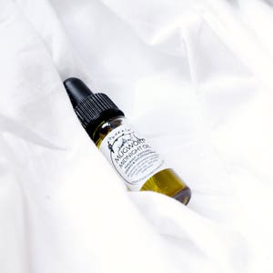 Pode incluir: Um pequeno frasco de vidro com um conta-gotas preto na parte superior cheio de um l&iacute;quido amarelo. O r&oacute;tulo diz "Mugwort Midnight Oil" e o descreve como um &oacute;leo arom&aacute;tico, calmante, purificante e m&aacute;gico de intui&ccedil;&atilde;o. O frasco est&aacute; sobre um fundo de tecido branco.