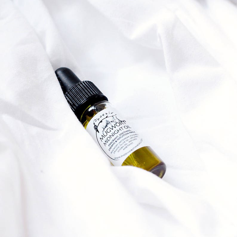 Pode incluir: Um pequeno frasco de vidro com um conta-gotas preto na parte superior cheio de um l&iacute;quido amarelo. O r&oacute;tulo diz "Mugwort Midnight Oil" e o descreve como um &oacute;leo arom&aacute;tico, calmante, purificante e m&aacute;gico de intui&ccedil;&atilde;o. O frasco est&aacute; sobre um fundo de tecido branco.