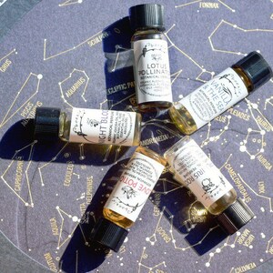 May include: Five small glass bottles with brown liquid and labels with names of different scents: Night Bloom, Love Potion, Iris Root, Vanilla & The Sea, and Lotus Pollinate. The bottles are arranged on a dark background with a constellation pattern.