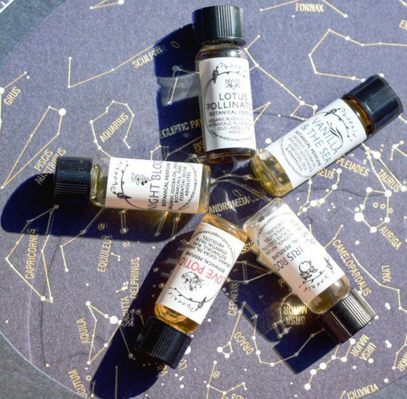May include: Five small glass bottles with brown liquid and labels with names of different scents: Night Bloom, Love Potion, Iris Root, Vanilla & The Sea, and Lotus Pollinate. The bottles are arranged on a dark background with a constellation pattern.