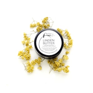 Op de afbeelding: Een klein zwart potje met een wit etiket waarop staat "Phoenix Linden Butter Skin Nourishing Fragrant Moisturizer Organic Shea Butter Organic Jojoba Oil Fresh Linden Flowers in Rounds of Enfleurage www.phoenixbotanical.com". Het potje is omringd door gedroogde gele bloemen.