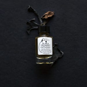 May include: A small glass bottle of black trumpet chthonic natural perfume. The bottle has a black cap and a white label with the text "Phoenir Black Trumpet Chthonic Natural Perfume Organic Cane Alcohol,  Black Trumpet Mushroom, Pure Plant Extracts, Essential Oils www.phoenirbotanicals.com".