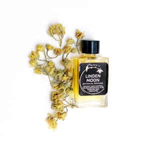 Peut inclure: Un petit flacon de parfum botanique Linden Moon en verre avec une &eacute;tiquette noire et un bouchon noir. L'&eacute;tiquette porte une inscription blanche qui dit "Phoenix Linden Moon Botanical Perfume Organic Cane Alcohol Infused Botanicals, Pure Essential Oils Absolutes www.phoenicanical.com". Des fleurs jaunes s&eacute;ch&eacute;es sont &eacute;parpill&eacute;es autour du flacon.