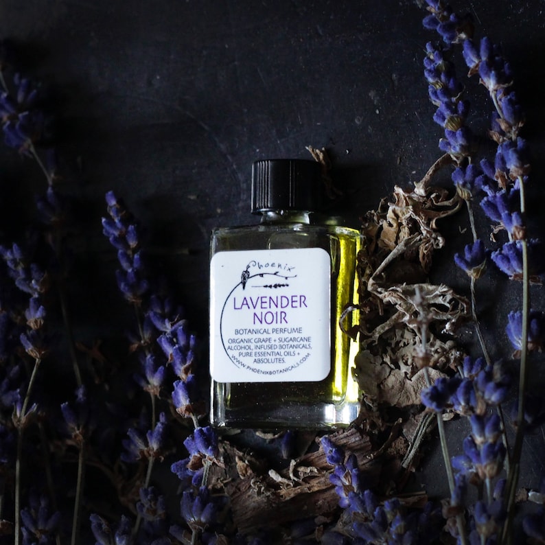 May include: A small glass bottle of Lavender Noir botanical perfume. The bottle has a black label with white text that reads "Phoenix Lavender Noir Botanical Perfume Organic Grape + Sugarcane Alcohol, Infused Botanicals Pure Essential Oils + Absolutes www.phoenixbotanicals.com".