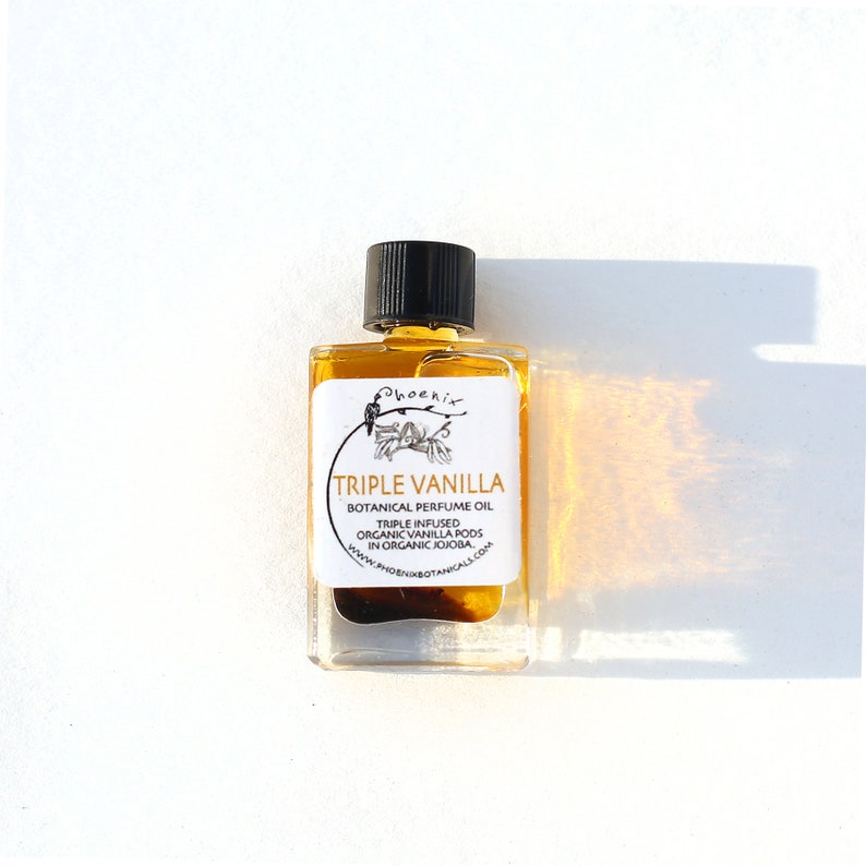 May include: A small, clear glass bottle with a black cap filled with a golden liquid. The bottle has a white label with black text that reads "Phoenix Triple Vanilla Botanical Perfume Oil Triple Infused Organic Vanilla Pods in Organic Jojoba www.phoenixbotanicals.com".
