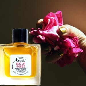 Pode incluir: Um frasco de perfume de vidro transparente com uma tampa preta e um rótulo branco que diz "Bed of Roses Botanical Perfume". A garrafa é preenchida com um líquido dourado. Uma mão segura uma rosa rosa ao lado da garrafa. O fundo é marrom escuro.
