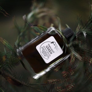 May include: A small glass bottle with a black cap containing a dark liquid. The bottle has a white label with the text "TWILIGHT GREEN BOTANICAL PERFUME" and a small star symbol.